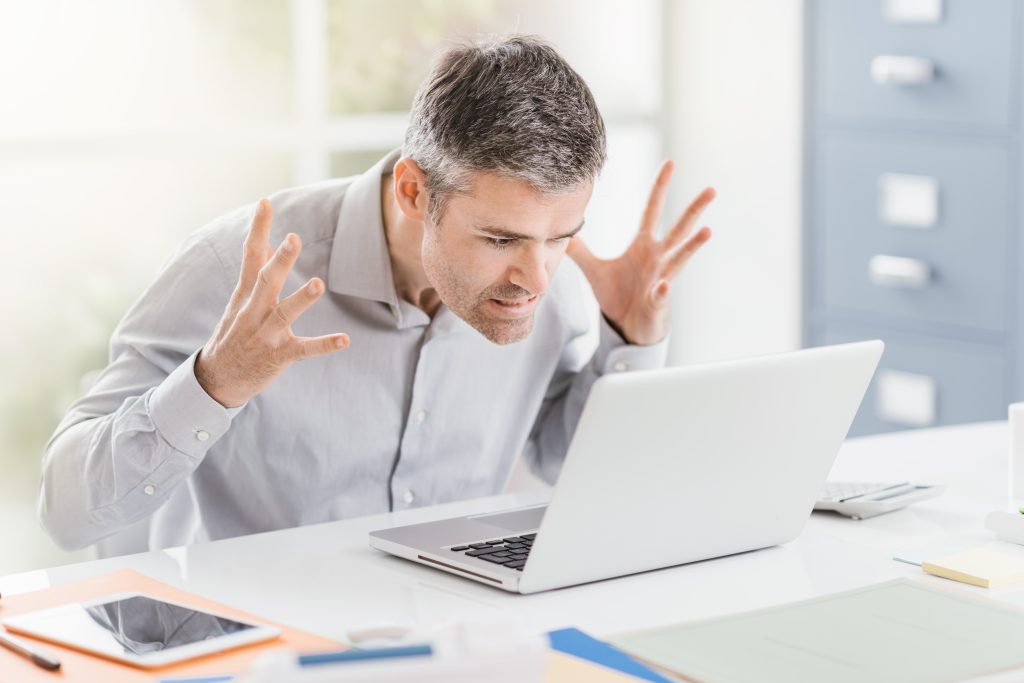 A man leaning forward with his hands up in frustration while staring at his laptop.