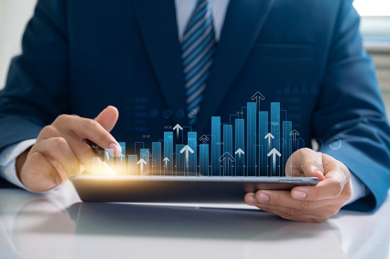 A man in a business suit using a tablet, holographic depictions of a bar graph and upward-pointing arrows appear from the screen.